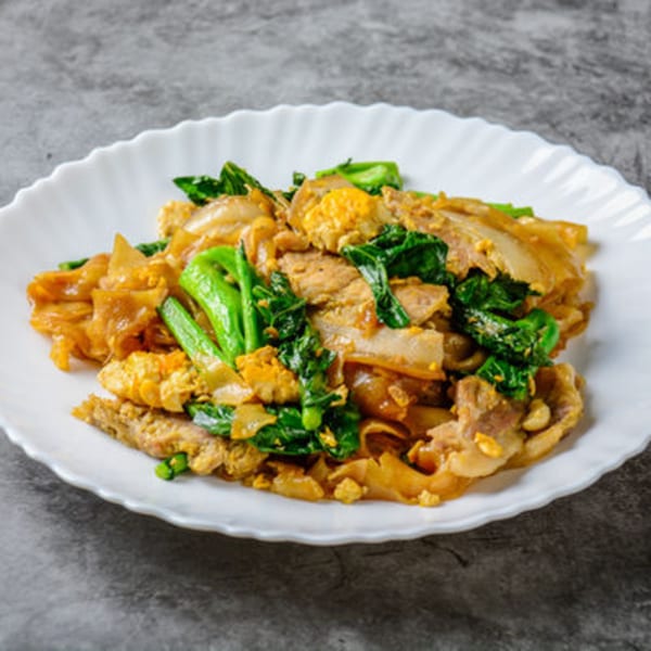 A white plate of Pad See Ew with wide noodles, pork, scrambled egg, and Chinese broccoli.