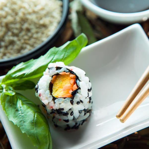 A single sushi roll with orange and yellow filling, rice, and black sesame seeds on a white plate with chopsticks.