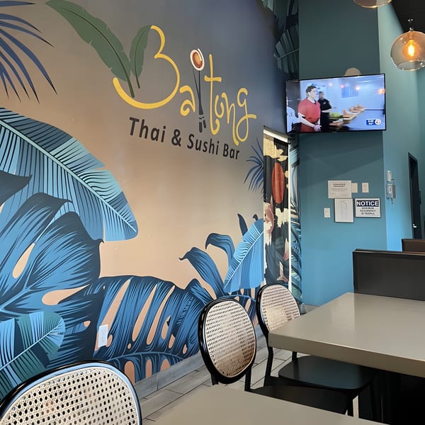 BAITONG Thai & Sushi Bar interior with a tropical leaf mural, tables, chairs, and a TV.