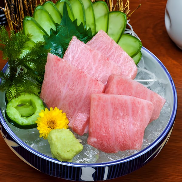 Pink tuna sashimi slices with green cucumber, wasabi, and a yellow flower on ice in a blue bowl.