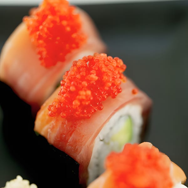 Close-up of three sushi rolls topped with bright orange tobiko, on a dark plate.