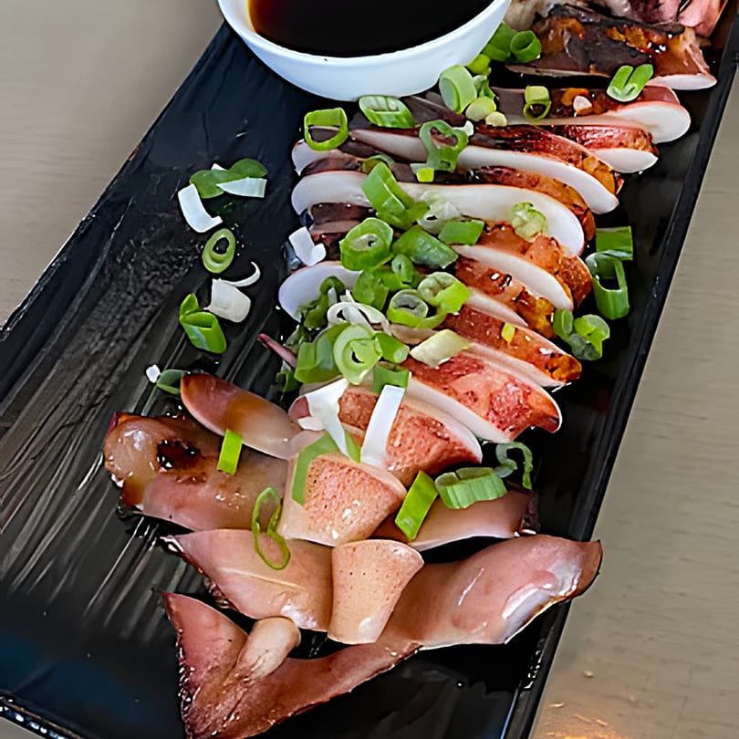 Grilled squid slices with green onions and a side of dark dipping sauce on a black plate.