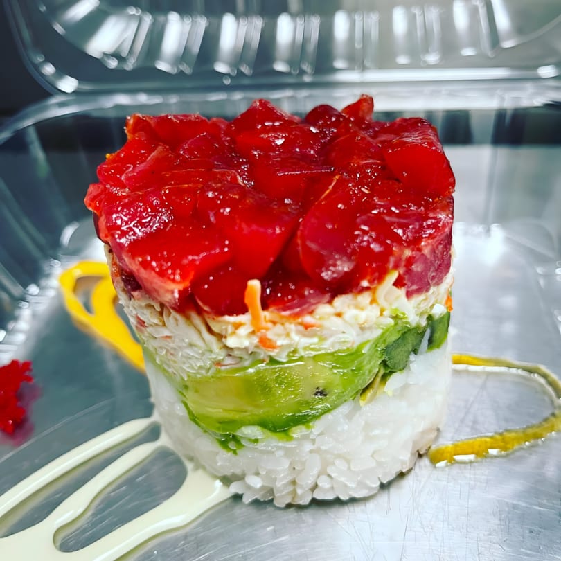 A vibrant sushi tower with layers of white rice, avocado, crab salad, and red tuna.