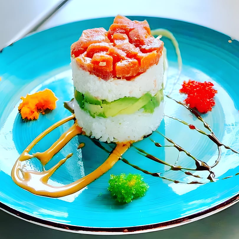 A vibrant sushi tower with salmon, rice, and avocado, garnished with roe and sauces on a blue plate.